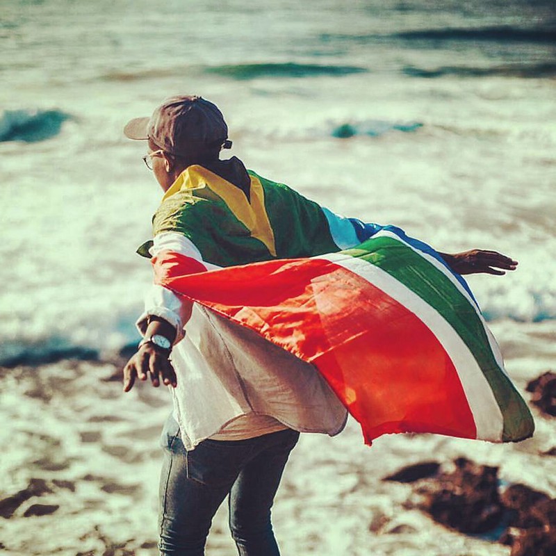 A young person running alongside the ocean with a South African flag draped over their back