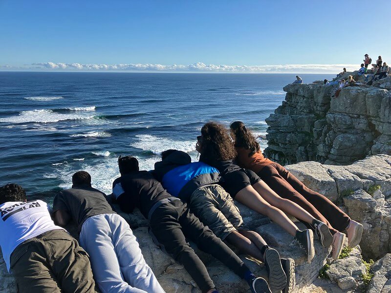 Tilting Futures gap year program alumni laying down side-by-side at the edge of a cliff overlooking the ocean