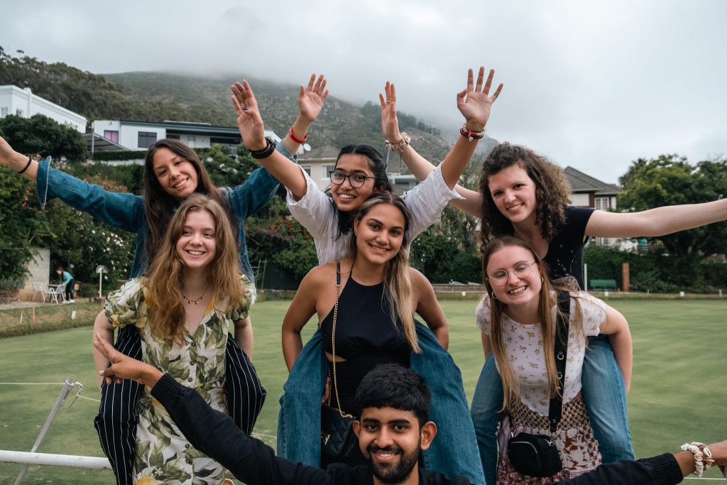 A group of seven young adults smiling and posing together outdoors, with some individuals sitting on others' shoulders. The background features a cloudy sky and green lawn, with buildings and trees visible in the distance.