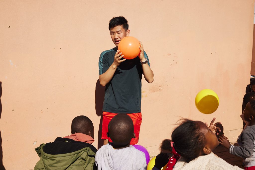 A young man holding an orange balloon while interacting with a group of children, some of whom are catching or reaching for colorful balloons. The scene is set against a pink wall.