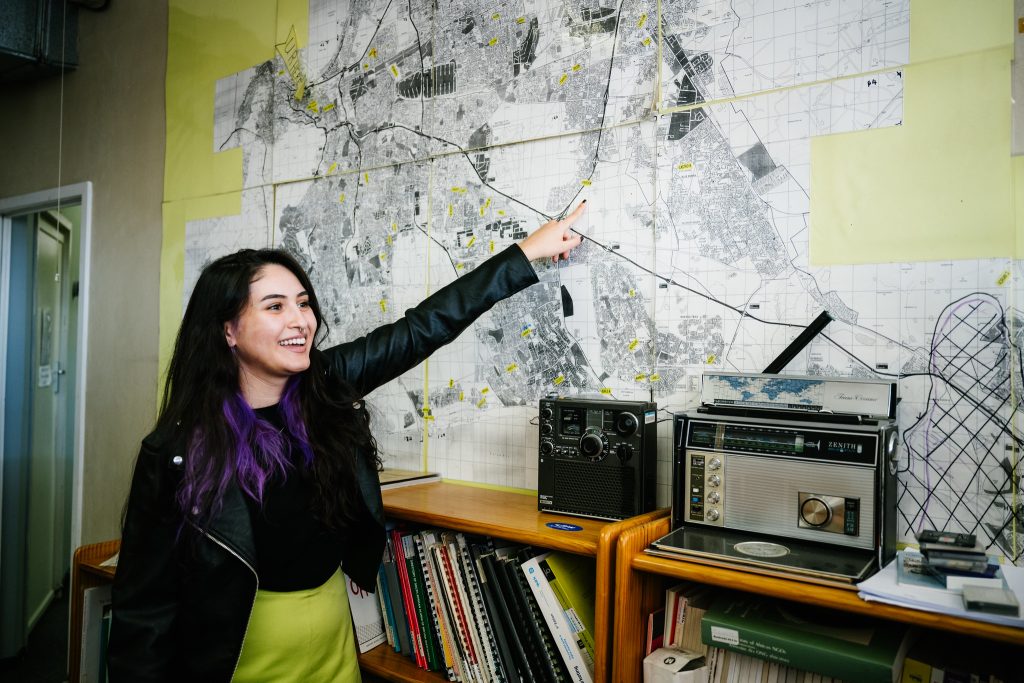 A person with long dark hair and purple highlights stands in front of a large map, pointing to a specific location. They are wearing a black leather jacket and a green skirt. On the table beside them are a vintage radio, a cassette player, and a shelf filled with books.