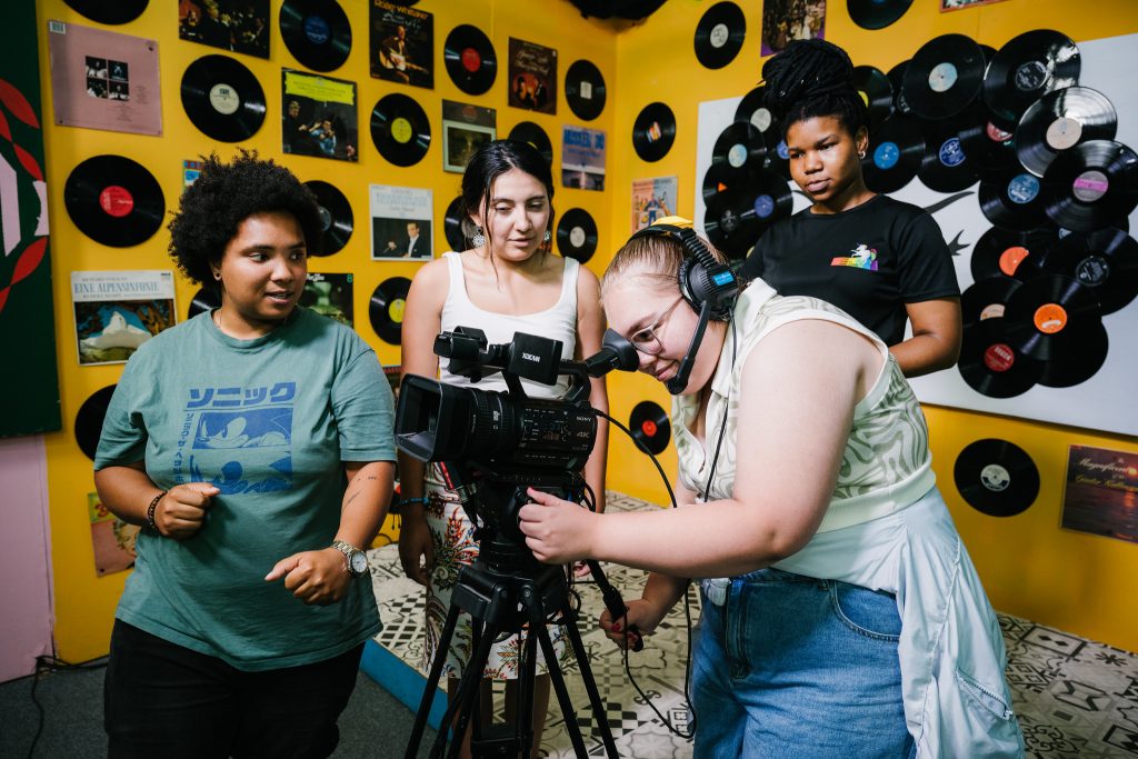 A group of four people in a music-themed room with vinyl records on the walls. One person, wearing a green Sonic T-shirt, is standing and looking towards another person, while two others are focused on a camera. The person operating the camera is wearing a headset and adjusting it, while the other person stands behind them offering assistance.