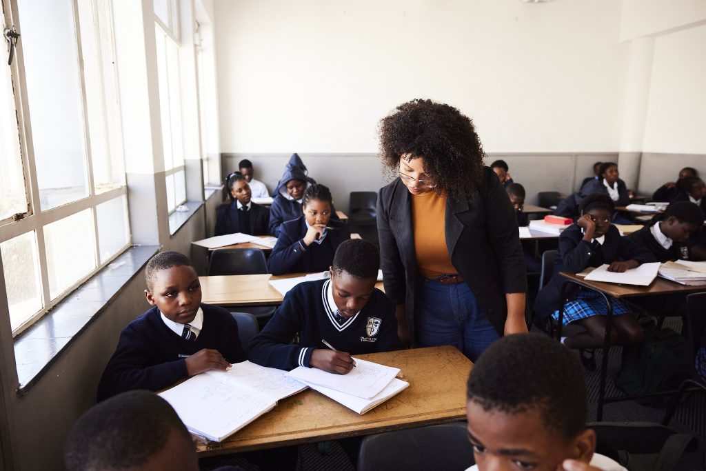 A teacher stands beside a student’s desk in a classroom, looking down at the student’s work while several other students in school uniforms sit at desks around the room, writing in notebooks.
