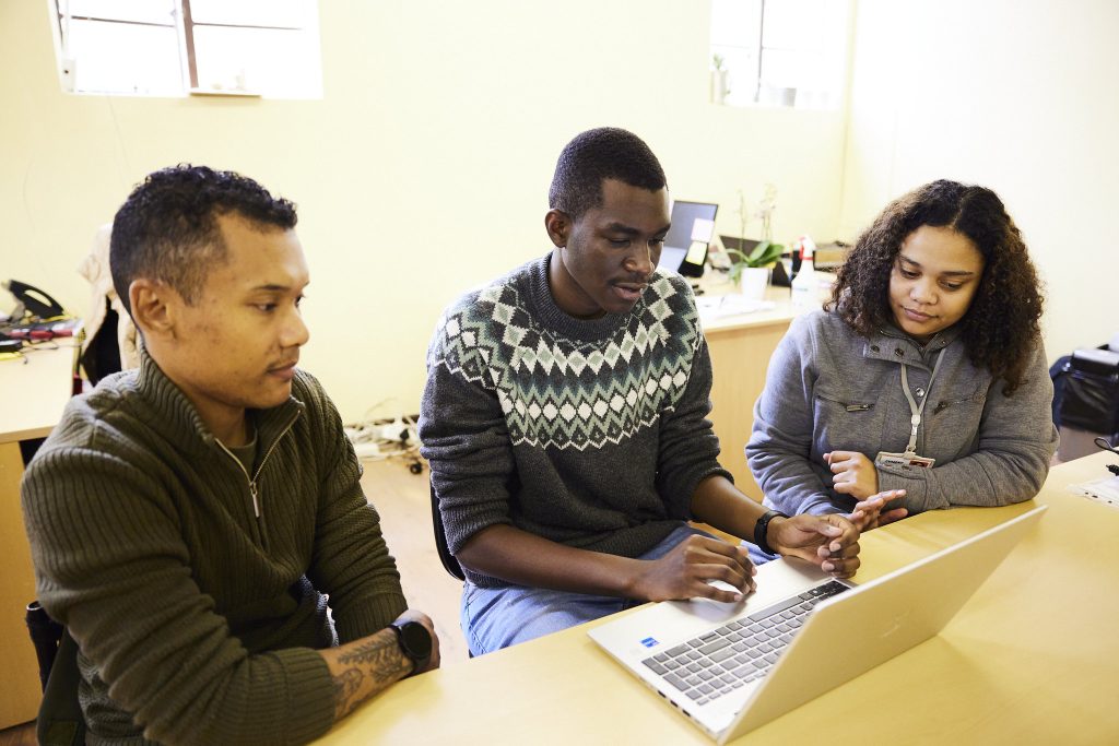 Three young adults sit together at a table, collaborating while looking at a laptop as one person gestures toward the screen and the others watch.