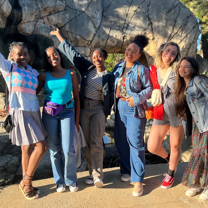 A group of six young women stand together outdoors in front of a large rock formation, smiling and posing with their arms around each other in bright sunlight.