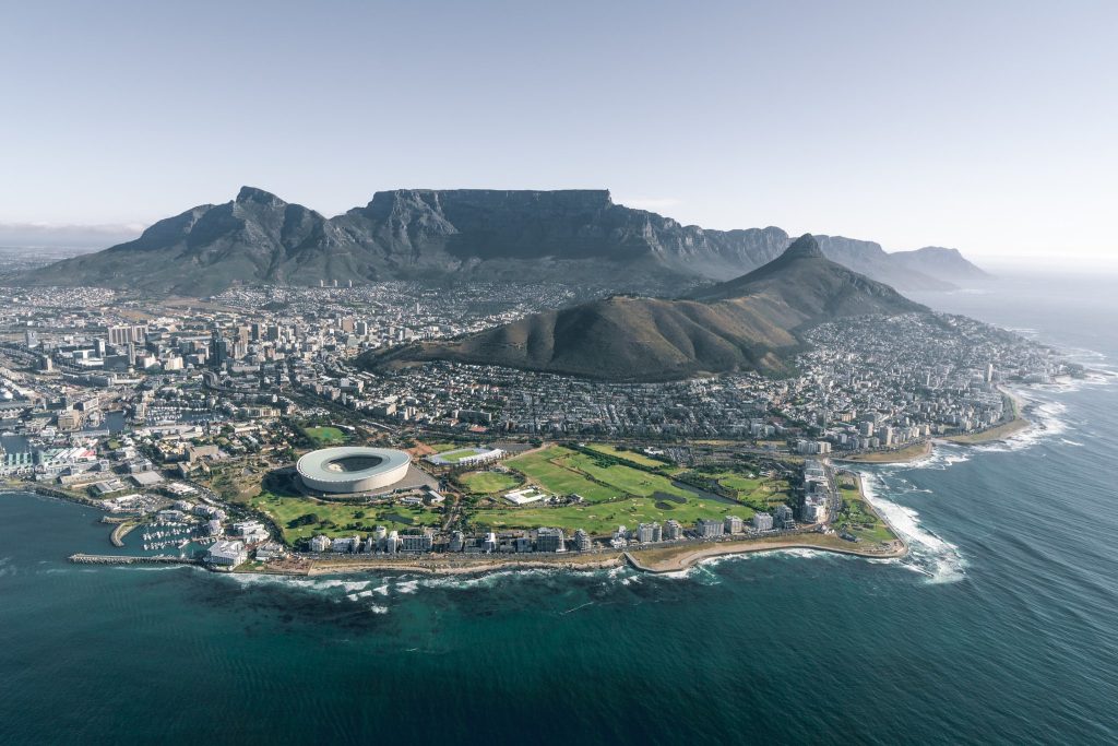 Aerial view of Cape Town, South Africa, showing the city's urban landscape nestled between the ocean and the iconic flat-topped Table Mountain. The V&A Waterfront and marina are visible on the left, DHL Newlands Stadium (now Cape Town Stadium) sits prominently near the shoreline, and residential and commercial districts spread across the foreground, with Lion's Head peak visible to the right of Table Mountain.