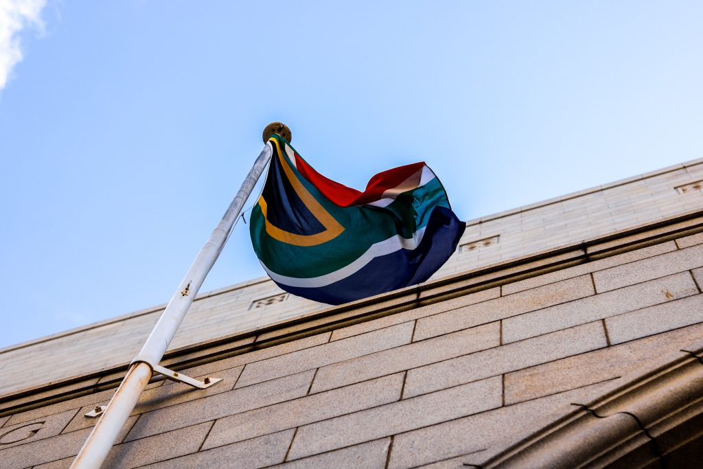 The South African flag flying from a white pole mounted on a stone building, photographed from below against a clear blue sky. The flag's distinctive colors — green, gold, black, white, red, and blue — are visible as it waves in the wind.