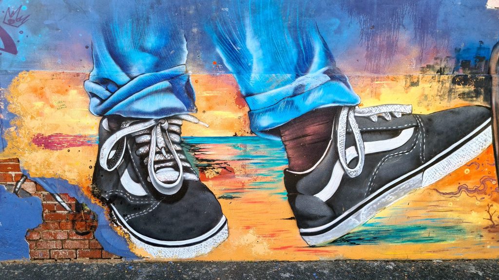 A vibrant street mural on a brick wall depicting a close-up, hyper-realistic painting of two black-and-white sneakers with loose laces, worn with rolled-up blue jeans. The colorful background features an abstract sunset landscape with warm oranges, yellows, and hints of teal suggesting water and sky.