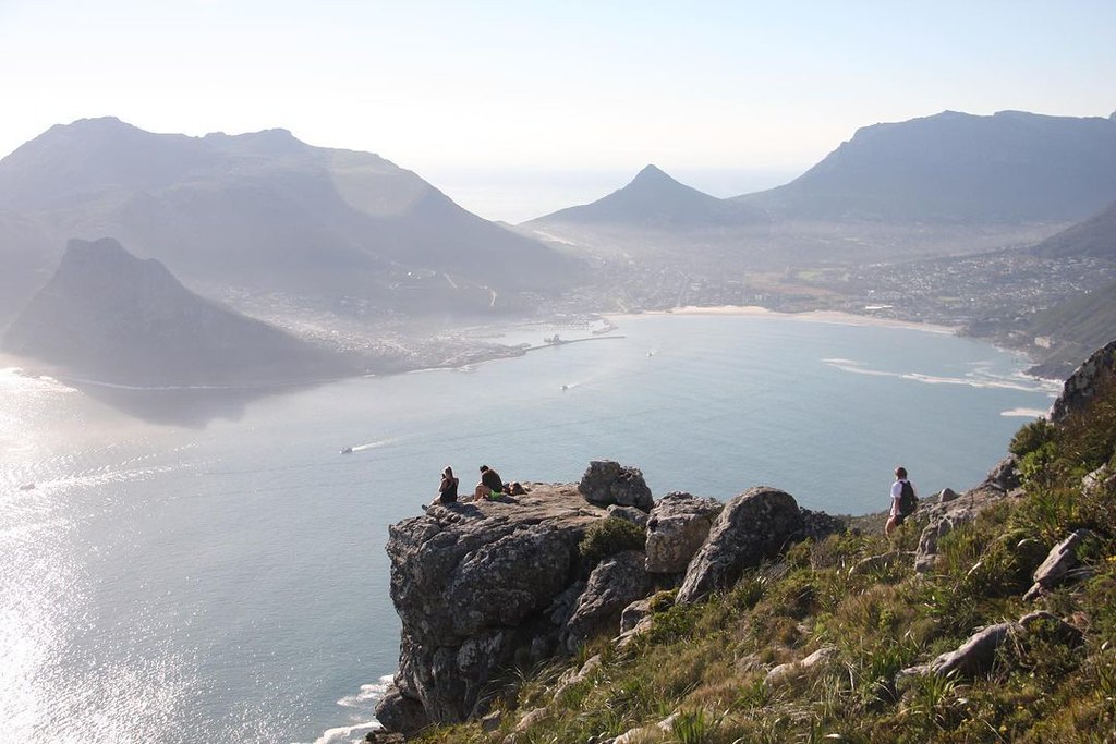 Hikers resting on a rocky cliff edge overlooking Hout Bay, a coastal town near Cape Town, South Africa, surrounded by dramatic mountain ranges. Two people sit on the rocky outcrop taking in the panoramic view of the bay and harbor below, while another hiker stands on the trail to the right. The scene is bathed in hazy sunlight with calm blue water and a sandy beach visible in the distance.
