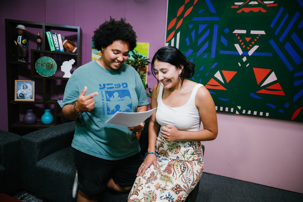Two young adults smile and review a sheet of paper together in a colorful room decorated with artwork and shelves.